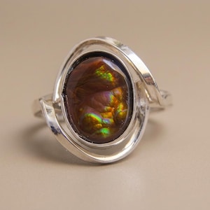 Puede incluir: Anillo de plata con una piedra preciosa ovalada en tonos marrones, naranjas y verdes. La piedra está engastada en una banda de plata en espiral, creando un diseño único y elegante. Esta es una pieza de joyería.