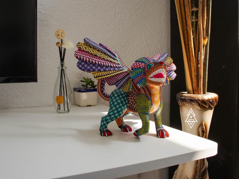 May include: A hand-painted wooden alebrije sculpture of a mythical creature with wings, a lion-like mane, and a wide-open mouth. The figure is decorated with intricate, colourful geometric patterns in shades of blue, yellow, orange, and purple.