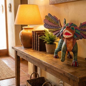 May include: A hand-painted wooden alebrije sculpture featuring a winged creature with a green body, orange chest, and multicoloured geometric patterns on its wings, displayed on a wooden console table next to a lamp, books, and a small potted plant.