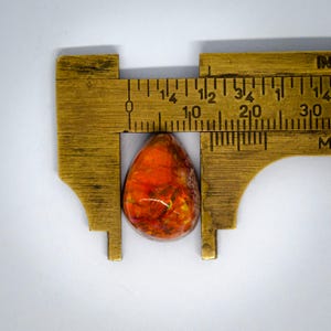 May include: A teardrop-shaped gemstone with vibrant orange and red hues, measured with a brass caliper. The stone's length is approximately 1 inch. The gemstone has a polished surface and displays a mix of colors.