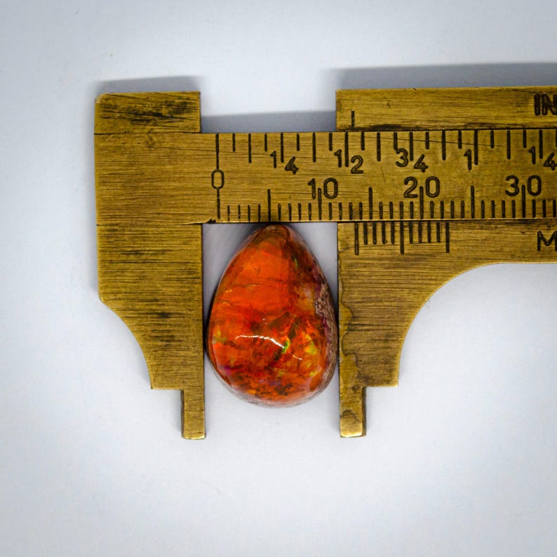 May include: A teardrop-shaped gemstone with vibrant orange and red hues, measured with a brass caliper. The stone's length is approximately 1 inch. The gemstone has a polished surface and displays a mix of colors.