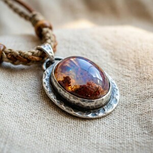 May include: A necklace featuring an oval-shaped, polished amber-colored stone set in a hammered silver-toned metal pendant. The pendant is attached to a braided tan cord necklace accented with small, round, dark brown wooden beads.