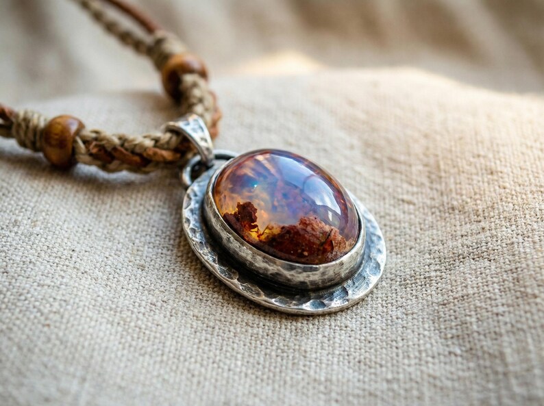 May include: A necklace featuring an oval-shaped, polished amber-colored stone set in a hammered silver-toned metal pendant. The pendant is attached to a braided tan cord necklace accented with small, round, dark brown wooden beads.