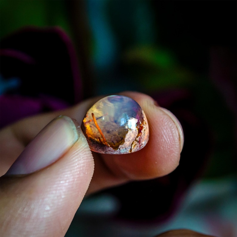 May include: A small, polished cabochon gemstone featuring a translucent, dome-shaped top with iridescent flashes of blue and white, transitioning into a textured, earthy base with warm orange and brown inclusions visible within the stone.
