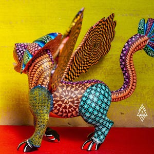 May include: A hand-painted alebrije sculpture of a mythical creature with wings, featuring intricate geometric patterns in vibrant colours including yellow, teal, orange, and purple. The figure has a long, curved tail and clawed feet.
