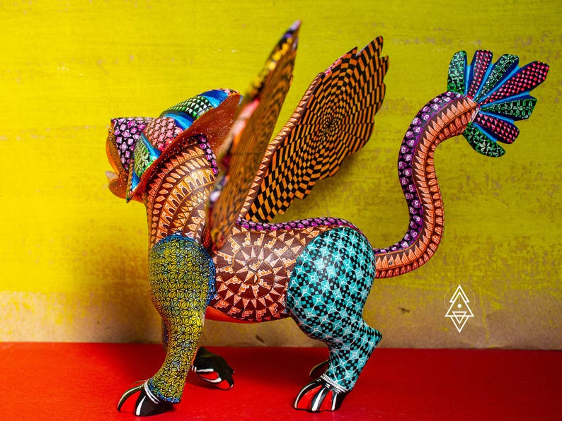 May include: A hand-painted alebrije sculpture of a mythical creature with wings, featuring intricate geometric patterns in vibrant colours including yellow, teal, orange, and purple. The figure has a long, curved tail and clawed feet.