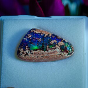 May include: A blue, green, and brown opal gemstone with a rough, natural shape. The opal is displayed on a white background.