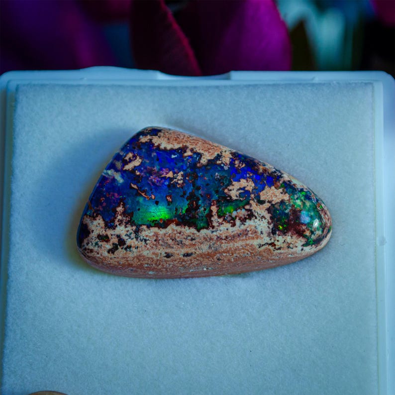 May include: A blue, green, and brown opal gemstone with a rough, natural shape. The opal is displayed on a white background.