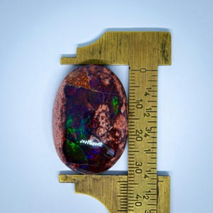 May include: A close-up of a colourful opal cabochon, measuring approximately 2.5 cm in length, with a red, brown, green, and purple colour pattern. The opal is being measured with a ruler.