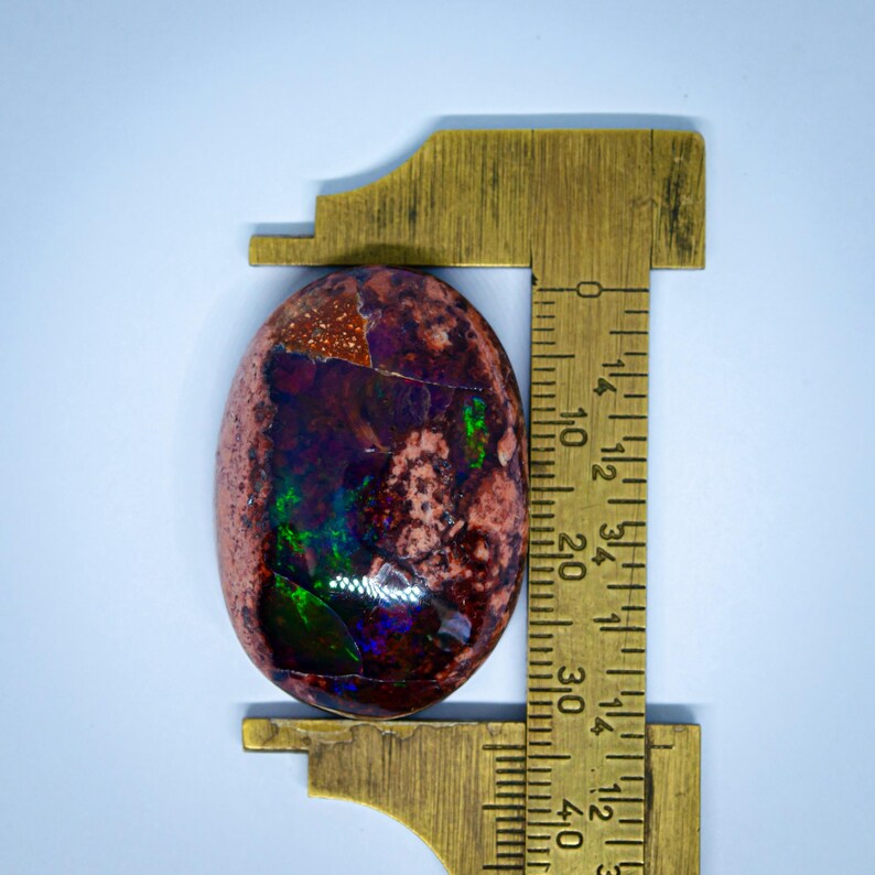 May include: A close-up of a colourful opal cabochon, measuring approximately 2.5 cm in length, with a red, brown, green, and purple colour pattern. The opal is being measured with a ruler.