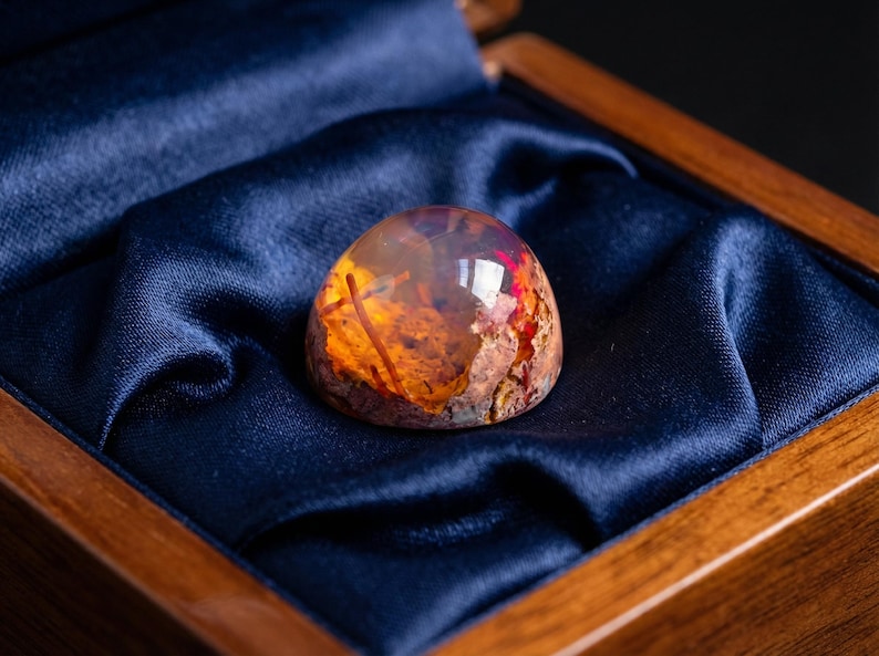 May include: A polished, dome-shaped cabochon gemstone featuring a translucent, amber-colored resin body with embedded organic inclusions, including reddish-brown fibrous elements and textured, earthy, multi-colored mineral-like fragments.