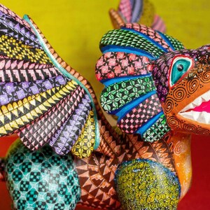 May include: A hand-painted wooden alebrije sculpture featuring a mythical creature with large wings, a lion-like head, and an open mouth showing white teeth. The body is covered in intricate geometric patterns in vibrant colours including yellow, purple, blue, green, and orange.