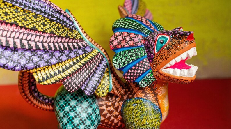 May include: A hand-painted wooden alebrije sculpture featuring a mythical creature with large wings, a lion-like head, and an open mouth showing white teeth. The body is covered in intricate geometric patterns in vibrant colours including yellow, purple, blue, green, and orange.
