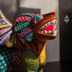 May include: A hand-painted wooden alebrije sculpture of a mythical creature with wings, featuring intricate geometric patterns in vibrant colours including orange, green, purple, and black. The creature has an open mouth with white teeth.