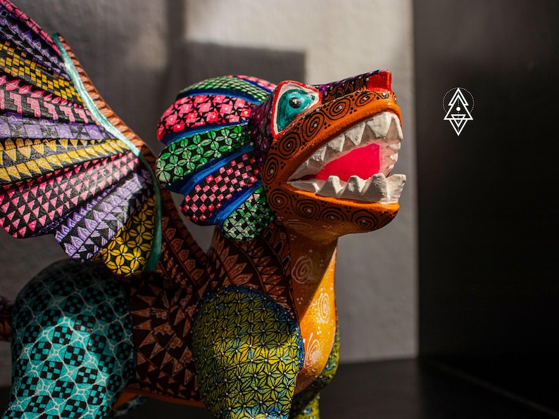 May include: A hand-painted wooden alebrije sculpture of a mythical creature with wings, featuring intricate geometric patterns in vibrant colours including orange, green, purple, and black. The creature has an open mouth with white teeth.