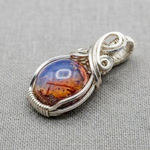 May include: A silver wire-wrapped pendant featuring a smooth, oval-shaped cabochon stone. The stone displays a gradient of deep blue at the top transitioning into a textured, mottled orange and brown base with visible dark needle-like inclusions.