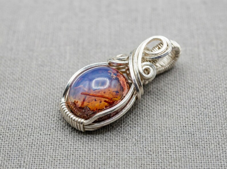 May include: A silver wire-wrapped pendant featuring a smooth, oval-shaped cabochon stone. The stone displays a gradient of deep blue at the top transitioning into a textured, mottled orange and brown base with visible dark needle-like inclusions.