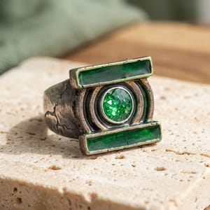 Puede incluir: Un anillo de tono plateado con una piedra preciosa verde circular central rodeada por un marco metálico con dos acentos rectangulares verdes horizontales. La banda tiene un acabado desgastado y texturizado con grietas.