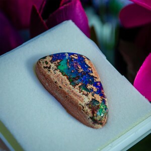 May include: A blue, green, and brown opal gemstone with a rough, natural finish. The opal is displayed on a white background.