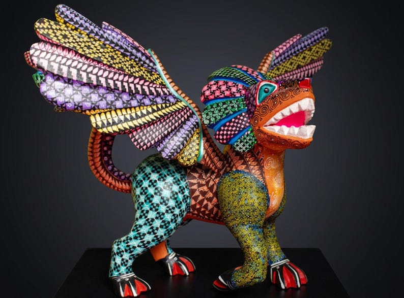 May include: A hand-painted alebrije sculpture of a mythical creature with wings, featuring a vibrant colour palette of orange, teal, yellow, and purple. The body is covered in intricate geometric patterns, including diamonds, swirls, and dots, with a wide-open mouth showing white teeth.
