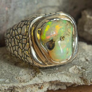 Puede incluir: Un anillo de plata con una gran piedra de ópalo ovalada. El ópalo muestra un juego de colores con tonos verdes, amarillos y anaranjados. La banda del anillo tiene un diseño texturizado en forma de escamas, añadiendo una estética única.