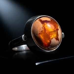 Puede incluir: Un anillo de plata con una gema grande y ovalada. La gema presenta tonos cálidos y terrosos con patrones anaranjados y rojos. El anillo se destaca sobre un fondo oscuro, resaltando los colores vibrantes de la piedra.