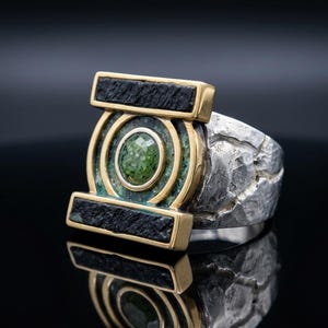 Puede incluir: Un anillo de plata con un marco dorado y un centro de piedra verde. El anillo tiene una banda texturizada y un diseño rectangular con detalles en negro. El anillo se muestra sobre una superficie negra reflectante.
