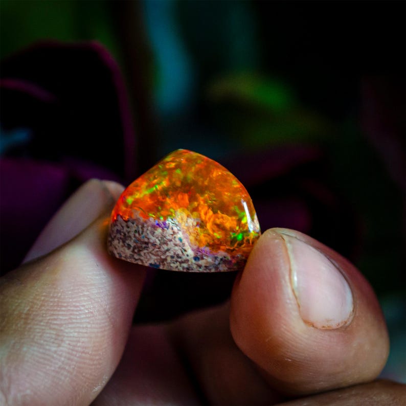 May include: A triangular-shaped opal gemstone with a vibrant display of orange, yellow, and green colors. The gemstone has a rough, textured base and a polished, iridescent top, showcasing a play of light.