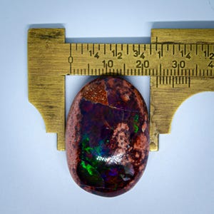 May include: A close-up of a dark red, brown, and green opal cabochon with iridescent flashes of colour. The opal is measured with a ruler, showing it is approximately 2.5 cm in diameter.
