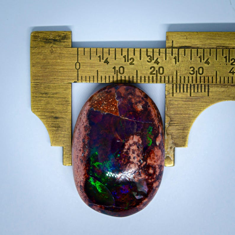 May include: A close-up of a dark red, brown, and green opal cabochon with iridescent flashes of colour. The opal is measured with a ruler, showing it is approximately 2.5 cm in diameter.