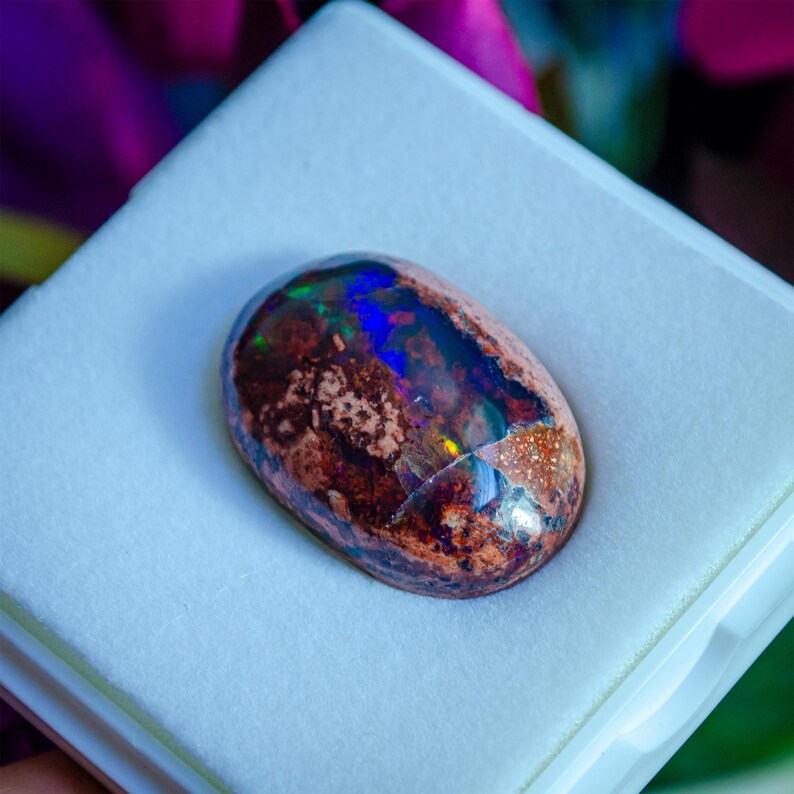 May include: A large, oval-shaped opal gemstone with a brown, red, and black base. The opal has a strong iridescent sheen with flashes of blue, green, and purple.