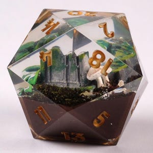 May include: A clear, multi-faceted D20 die with a miniature landscape inside. The scene includes a castle, mushrooms, and greenery set in a brown base. Gold numbers mark the die's faces, creating a unique tabletop gaming accessory.