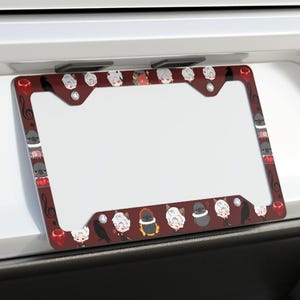 Könnte beinhalten: Ein rechteckiger Kennzeichenhalter mit dunkelrotem Hintergrund, der ein sich wiederholendes Muster aus kleinen, stilisierten Zeichentrickfiguren zeigt, darunter weiße Schafe, schwarze Vögel und rote Musiknoten, montiert an einem weißen Fahrzeug.