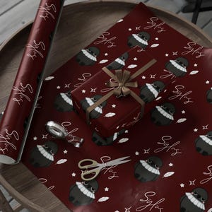 May include: A roll of burgundy wrapping paper and a wrapped gift box with a repeating pattern of a cartoon bird with a white collar, stars, and feathers. The gift is tied with a brown ribbon. Gold scissors and tape are also on the wooden table.