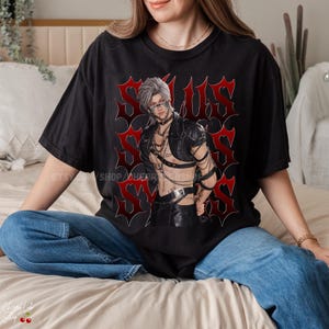 Può includere: T-shirt nero con grafica di una figura stilizzata con capelli argentati, che indossa un'imbracatura in pelle e catene. La grafica è ambientata su uno sfondo rosso in stile gotico con la parola "SOULUS" ripetuta. La maglietta è indossata da una persona in jeans blu.