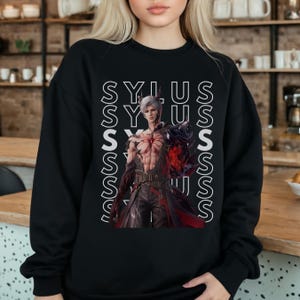 K&ouml;nnte beinhalten: Ein schwarzes Langarm-Sweatshirt mit einem Grafikdruck eines m&auml;nnlichen Charakters mit wei&szlig;em Haar und dunkler R&uuml;stung. Der Name SYLUS ist in wei&szlig;en vertikalen Textspalten hinter dem Charakter wiederholt, der ein rotes Emblem tr&auml;gt.