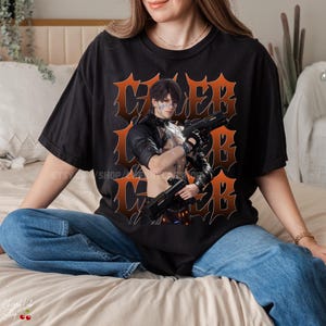 Può includere: T-shirt nera con grafica arancione di una persona che tiene due pistole, con la parola "CYBER" in caratteri stilizzati. Il design ha un aspetto vintage e usurato. La maglietta è di vestibilità ampia.
