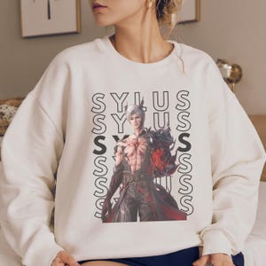 K&ouml;nnte beinhalten: Ein cremefarbenes Sweatshirt mit Rundhalsausschnitt und einem Grafikdruck eines m&auml;nnlichen Charakters mit wei&szlig;em Haar und muskul&ouml;sem Oberk&ouml;rper. Der Charakter tr&auml;gt dunkle, geschichtete Kleidung mit roten Akzenten. Der Text SYLUS ist vertikal wiederholt.