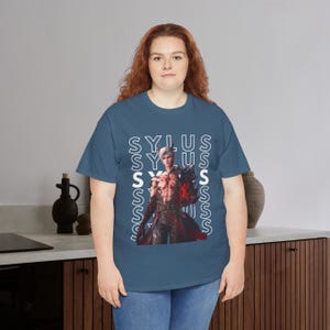 K&ouml;nnte beinhalten: Ein blaues T-Shirt mit kurzen &Auml;rmeln und einem Grafikdruck eines Charakters mit wei&szlig;em Haar und einem rot leuchtenden Arm. Der Name SYLUS wird in wei&szlig;en vertikalen Textspalten hinter dem Charakter wiederholt, was ein geschichtetes Design erzeugt.