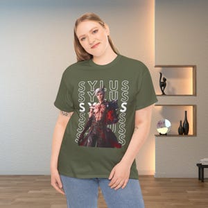 K&ouml;nnte beinhalten: Ein olivgr&uuml;nes T-Shirt mit einem Grafikdruck eines m&auml;nnlichen Charakters mit wei&szlig;em Haar und dunkler Kleidung. Das Wort SYLUS ist in wei&szlig;em, vertikalem Text hinter dem Charakter wiederholt. Das T-Shirt hat kurze &Auml;rmel und einen Rundhalsausschnitt.