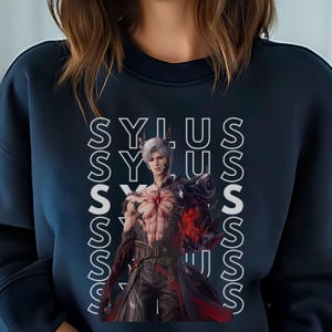 K&ouml;nnte beinhalten: Ein dunkelblaues Sweatshirt mit Rundhalsausschnitt und einem sich wiederholenden vertikalen wei&szlig;en Textmuster des Wortes SYLUS. Mittig auf der Vorderseite befindet sich eine Grafik eines silberhaarigen Charakters mit roten Markierungen.