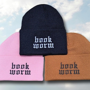 May include: Three beanies in different colors: pink, black, and brown. Each beanie has the words "book worm" embroidered on the front in a gothic font.