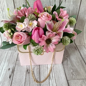 Artificial arrangement in pink and white in a bag with gold coloured detail and chain handles .
