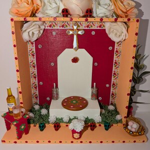 May include: A small wooden altar decorated with flowers, a cross, and miniature skulls. The altar is painted in shades of orange, red, and white. It features a small wooden stool with a bottle and a glass on top.