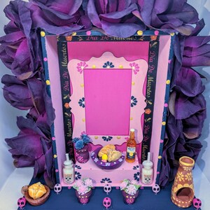 May include: A Day of the Dead altar with a pink and blue frame, adorned with purple flowers. It features miniature food, drinks, and decorative skulls. The altar includes the text "Dia De Muertos" on a ribbon.