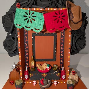 May include: A small wooden altar decorated with black roses, red and green paper flags, and a framed picture. The altar also includes a bottle of tequila, a bowl of bread, a glass of beer, and a few other decorative items.