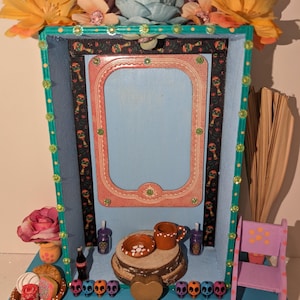 May include: A small wooden altar decorated with colorful flowers, a framed mirror, and a miniature chair. The altar also features a small heart, sugar skulls, and a clay cup and bowl on a wooden log.
