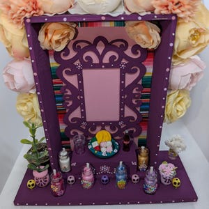 May include: A decorative purple shadow box with a pink interior and a decorative frame. The box is adorned with faux flowers and miniature items, including small bottles, a plate of food, and a miniature soda bottle. The background features a colorful striped fabric.