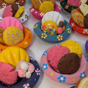 Może przedstawiać: Kolekcja małych, ręcznie malowanych ceramicznych misek z kolorowymi wzorami kwiatowymi, z kt&oacute;rych każda wypełniona jest miniaturowymi glinianymi replikami r&oacute;żnych wypiek&oacute;w, w tym ciastek, ciasteczek i chleba w odcieniach r&oacute;żu, ż&oacute;łci i brązu.