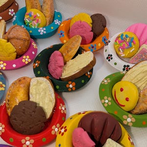 Może przedstawiać: Kolekcja miniaturowych misek pomalowanych na jasne kolory w białe kwiatowe wzory, z kt&oacute;rych każda zawiera małe gliniane produkty spożywcze, takie jak kromki chleba z pastą, ciasteczka, wypieki i kolorowe pączki z posypką.